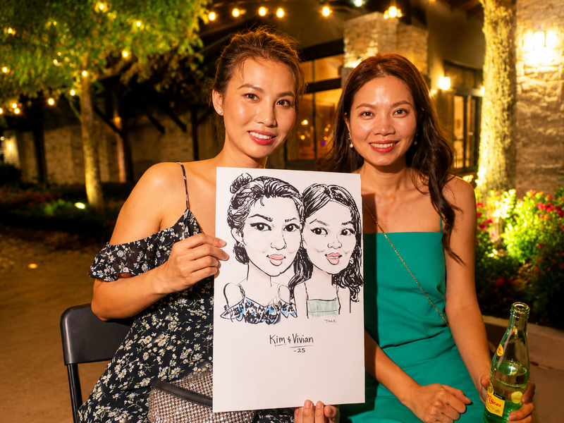 Two friends holding a duo caricature at an outdoor neighborhood evening event